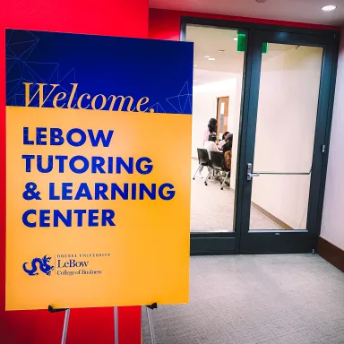 A blue and yellow sign with the text LeBow Tutoring and Learning Center, with a red wall and a glass window and door visible in background