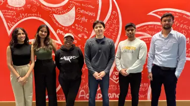Six college students standing against a bright red wall decorated with a dragon illustration and handwritten messages