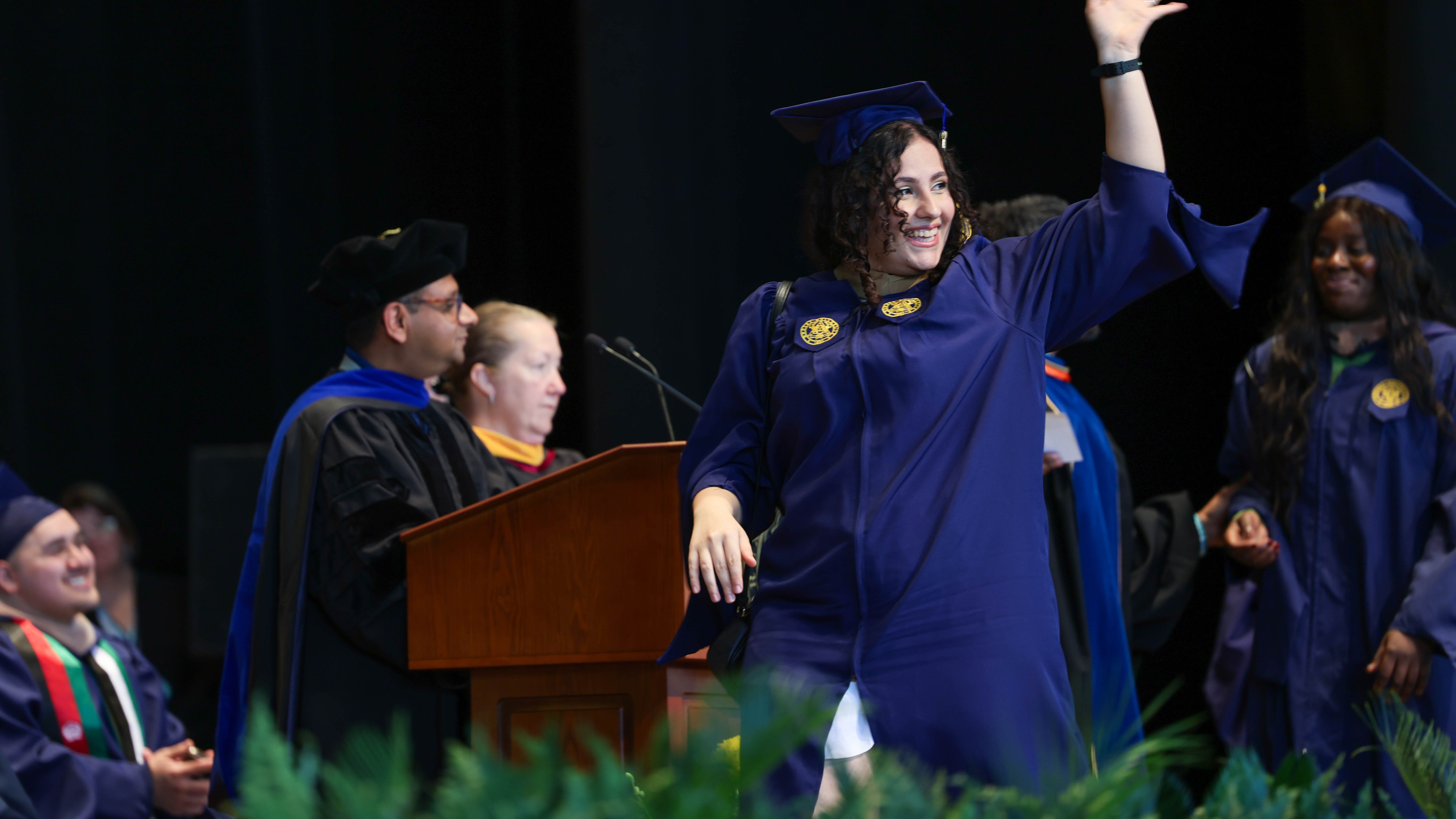 A young woman wearing a navy commencement gap and gown walks across the stage and waves with her left hand