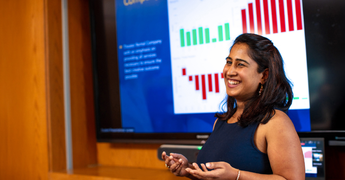 A graduate student making a business presentation in a boardroom with corporate logos visible in the background
