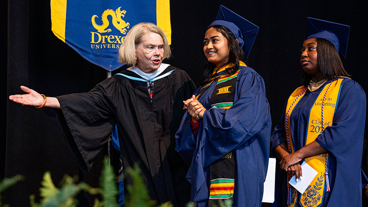 Two students in graduation robes being directed by a staff member with the Drexel University logo on a banner visible in the background