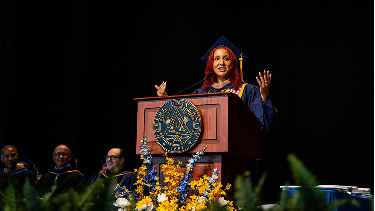 A young woman with red hair wearing a graduation robe and cap standing at a podium gesturing with both hands