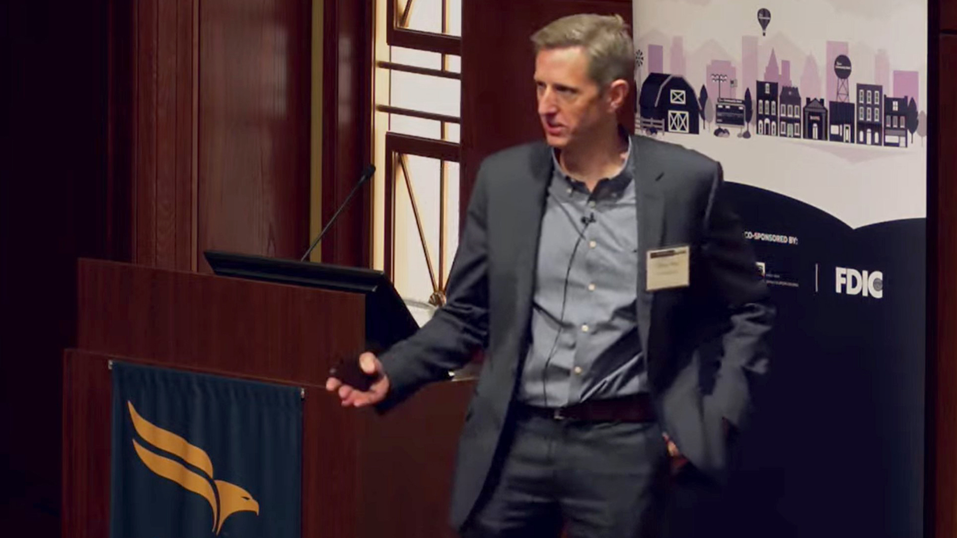 Professor Greg Nini leading discussion at the Community Banking Research Conference Man wearing blue suit and light blue dress shirt standing next to a podium with a computer screen and microphone visible