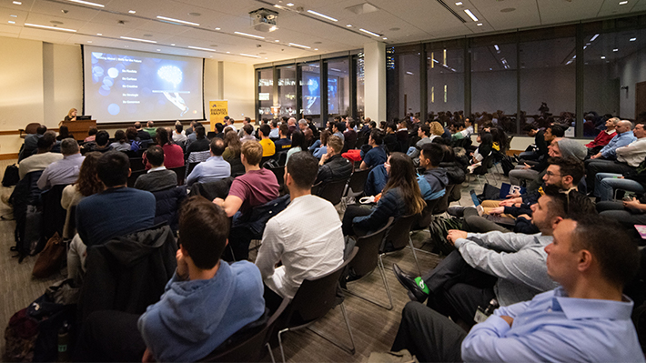 Crowd looks at screen and presenter in a large auditorium 