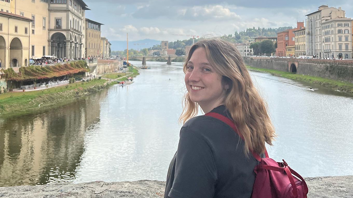 A young woman wearing a black shirt and red backpack standing on a bridge overlooking a river