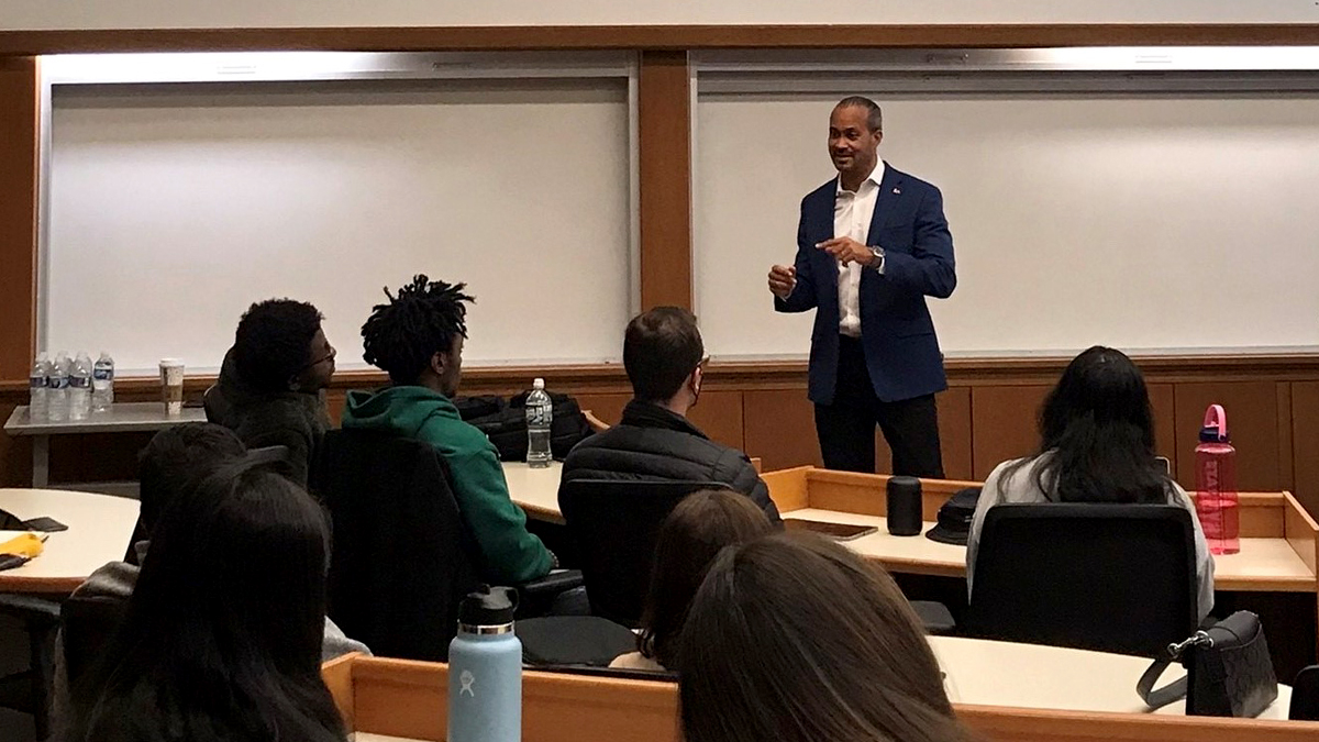 A man in a navy blue at the front of a classroom presenting to a group of students, the back of whose heads are visible