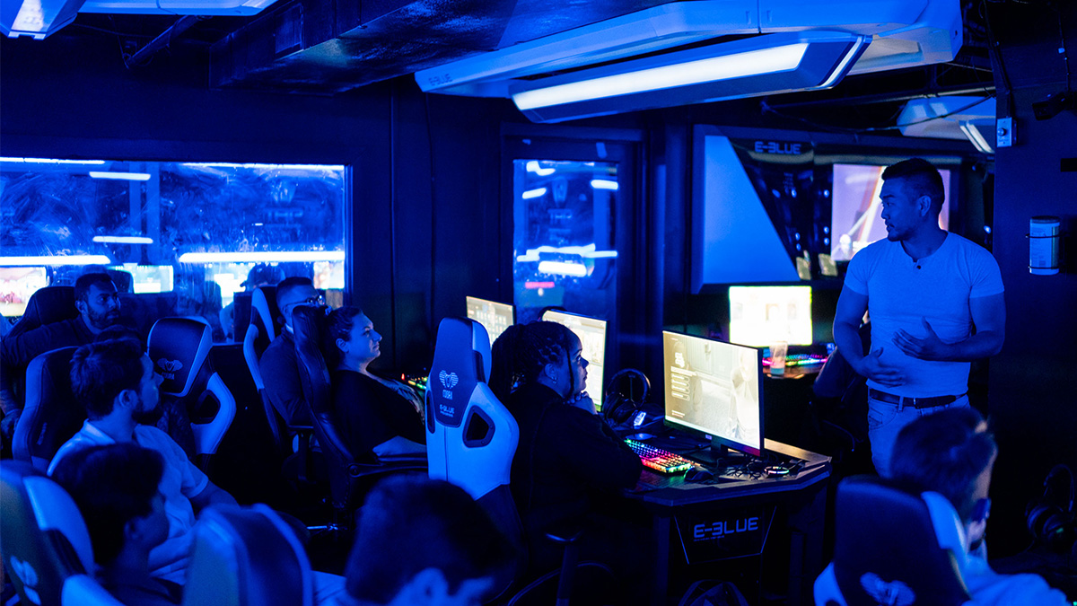 Several people seated at computer screens in a dark room with one man standing at the front