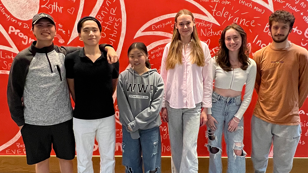 Six college students standing against a bright red wall decorated with a dragon illustration and handwritten messages