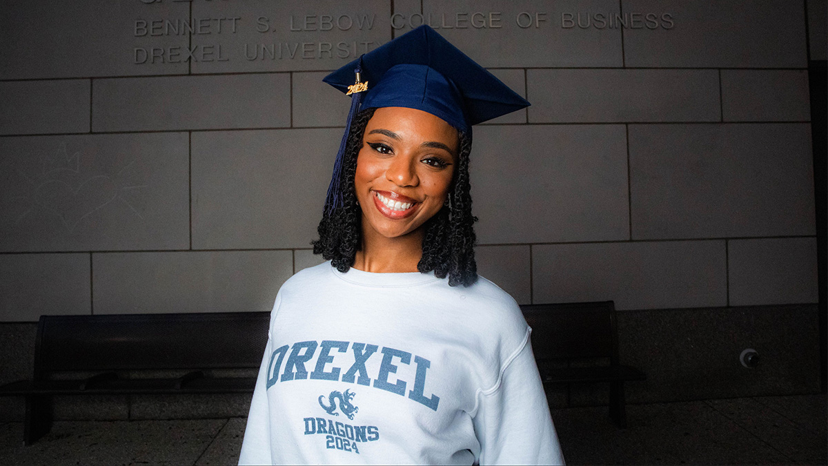 A young woman wearing a Drexel sweatshirt and a mortarboard posing against the exterior of Gerri C. LeBow Hall