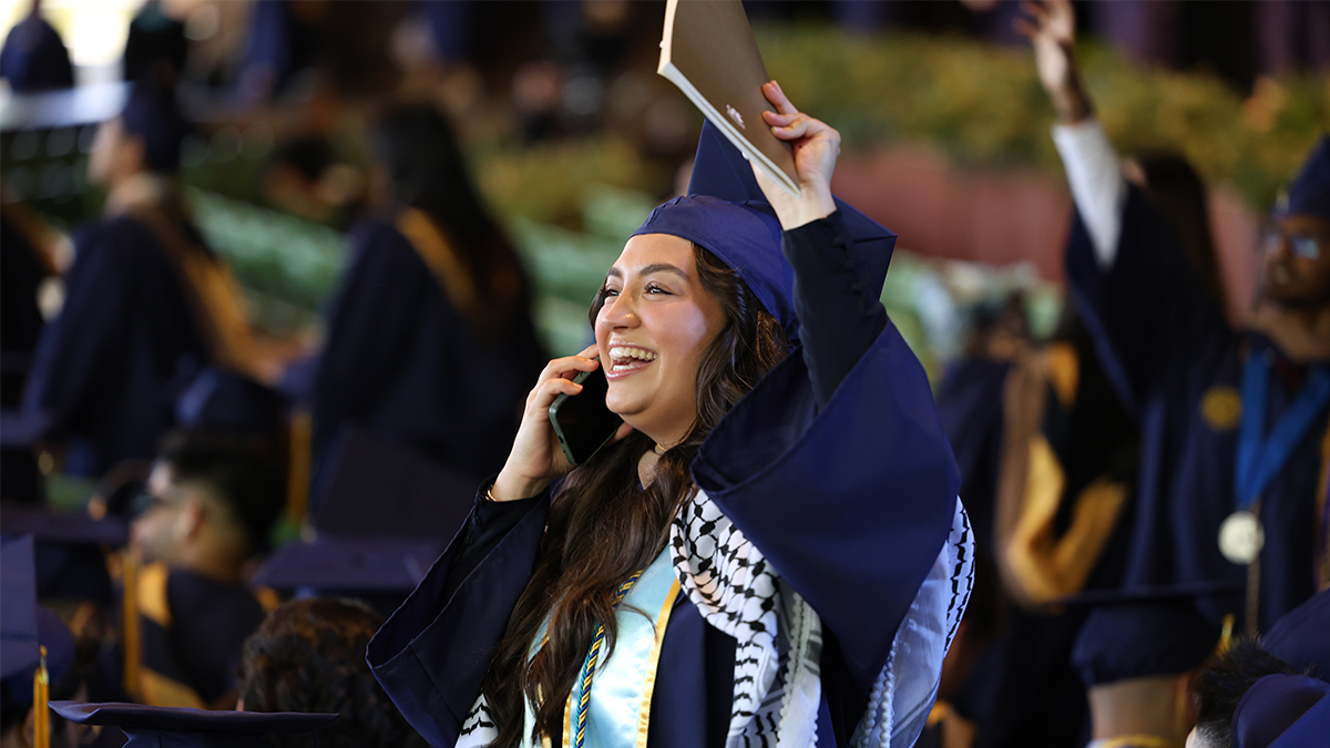 Aisa Feratovic, BS marketing '24 A female student in a blue graduation cap and gown waving to her family while talking on the phone