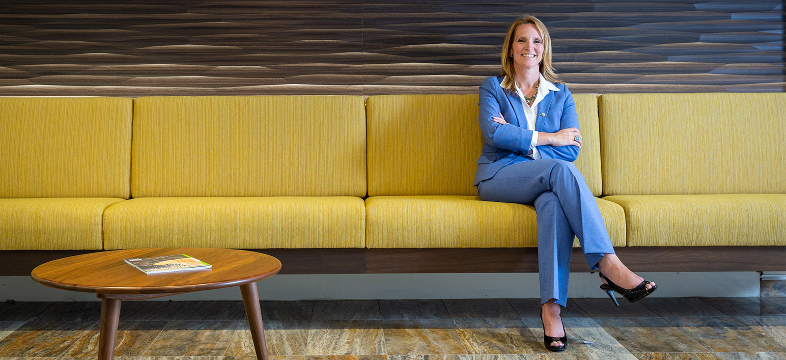 A woman in a light blue business suit seated on a yellow sofa with a short wooden table visible at left