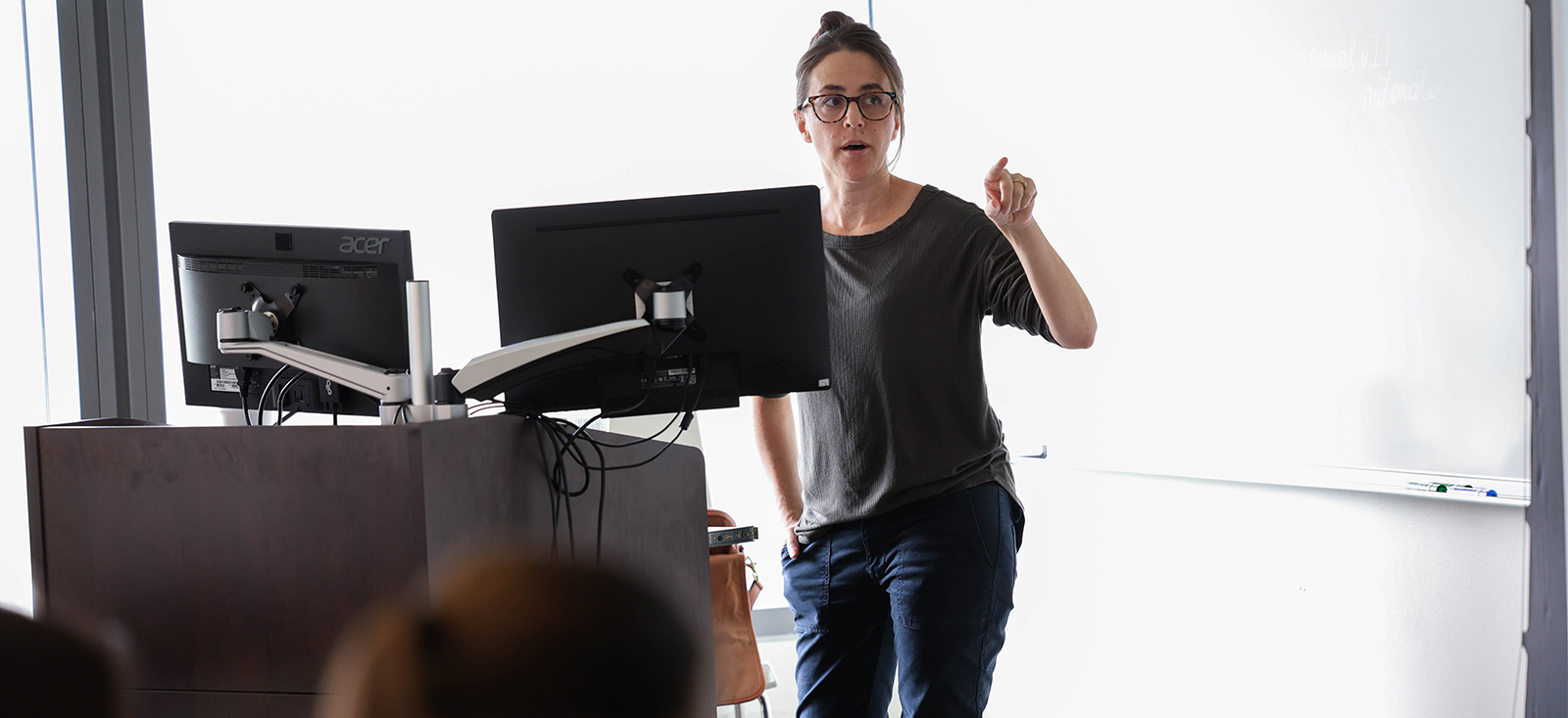 Anna Devlin, PhD, associate professor of decision sciences, teaching undergraduate students A woman with brown hair and glass pointing with her left hand and standing behind two computer monitors