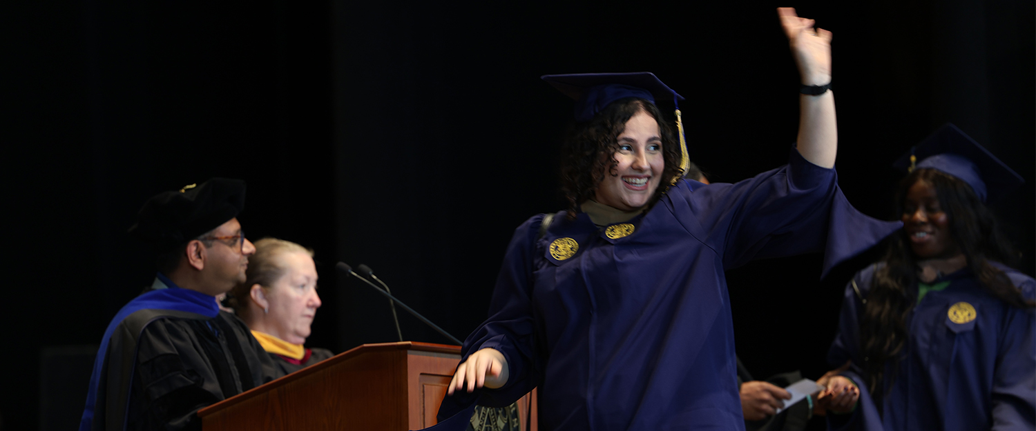 A young woman wearing a navy commencement gap and gown walks across the stage and waves with her left hand