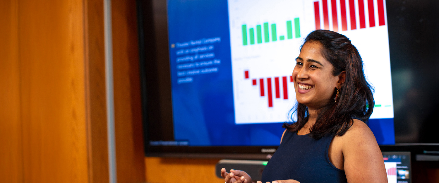 A graduate student making a business presentation in a boardroom with corporate logos visible in the background