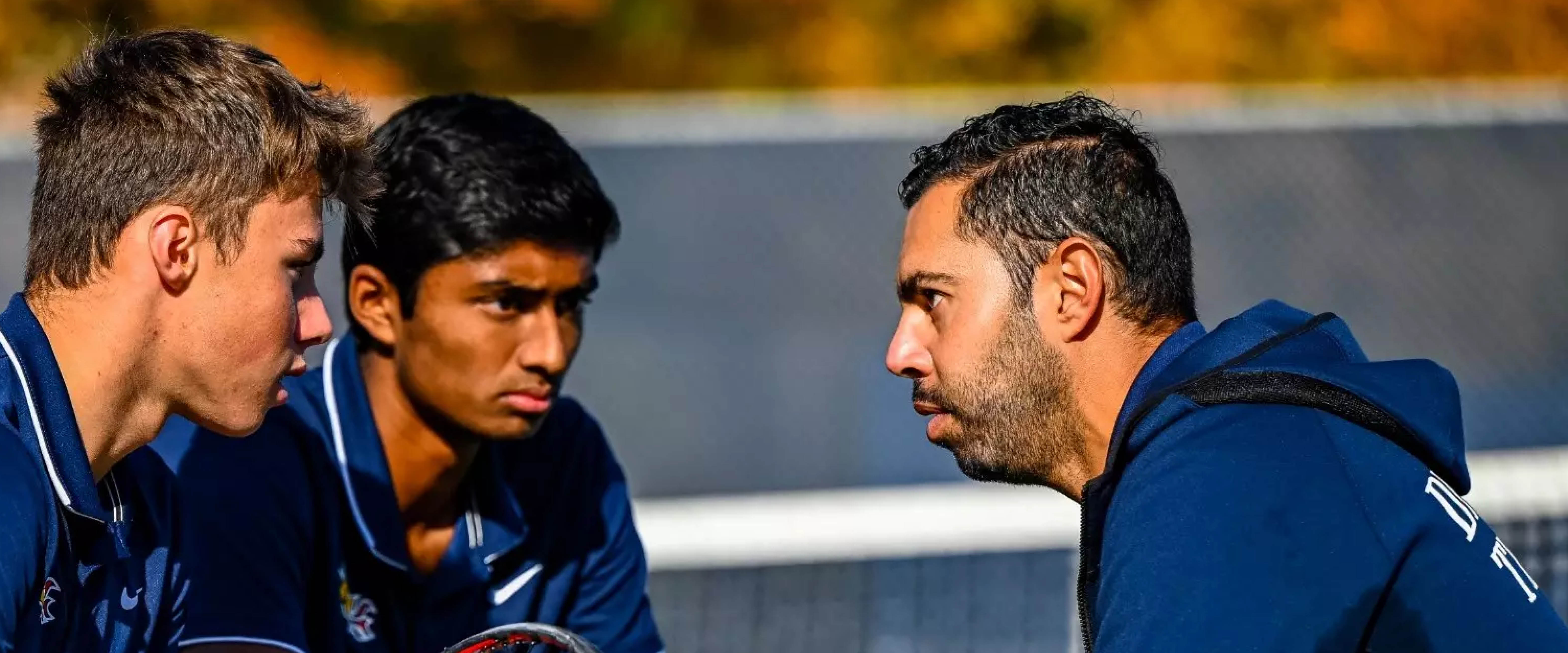 Two young men wearing navy polo shirts facing and talking to an older man wearing a navy jacket