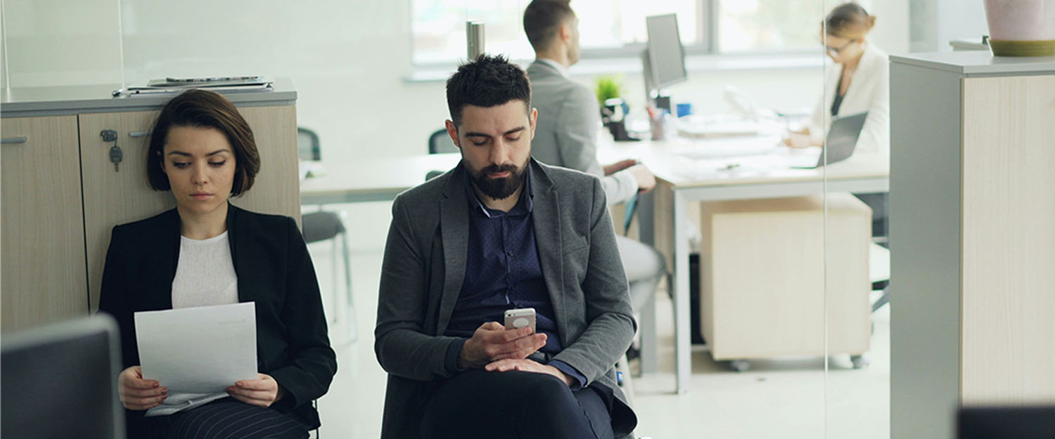A young woman holding a piece of paper and a young man holding a cell phone seated in an office wearing business attire