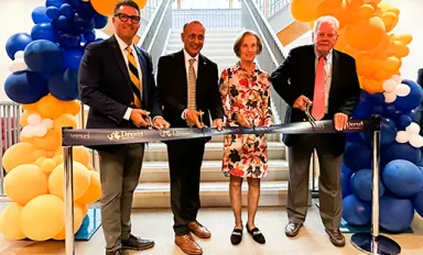 Four individuals holding scissors and cutting a ribbon suspended between two stanchions. Navy and gold balloons are visible behind and to the sides.