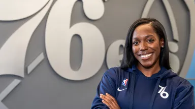Millie Watson, MS '19 A young Black woman in a blue sweatshirt with the Philadelphia 76ers logo, standing in front of a large silver rendition of the 76ers logo and with her arms crossed in front of her chest