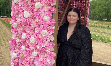 A young woman wearing a black dress with a floral print standing next to a long, vertical display of pink and white flowers