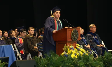 A young man wearing a navy commencement cap and gown standing a podium with the Drexel University seal on the front