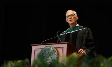 A man wearing glasses standing at a podium wearing a black graduation robe with a green stole
