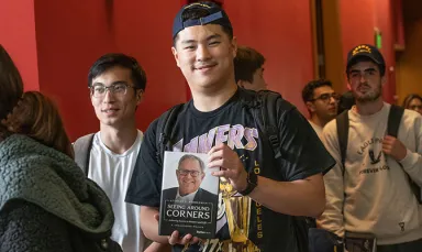 A young man wearing a backwards navy hat and a Los Angeles Lakers shirt holding a copy of Seeing Around Corners, a book by Stanley C. Middleman