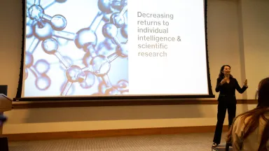 A woman presenting next to a large screen with the words Decreasing returns to individual intelligence & scientific research visible