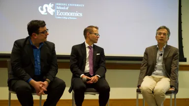 Three men seated, logo for Drexel School of Economics visible on screen in background