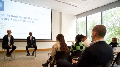 Two men in business suits sitting in chairs with young people seated visible to the right