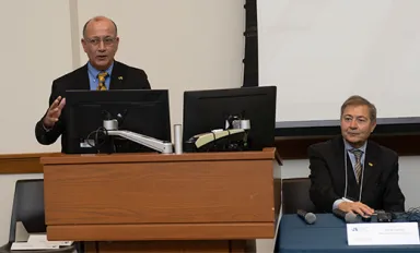 A man wearing glasses standing at a podium with a computer screen visible to the right, next to a man in a business suit seated