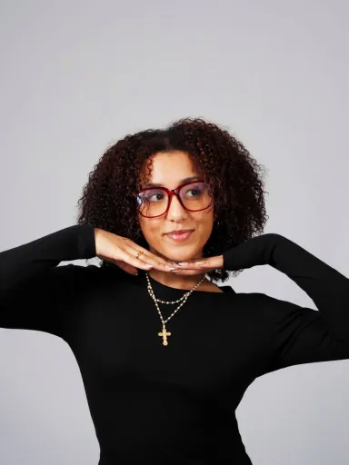A young woman wearing a black sweater, a necklace and glasses visible from the waist up