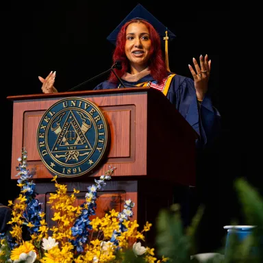 A young woman with red hair wearing a graduation robe and cap standing at a podium with the Drexel University seal