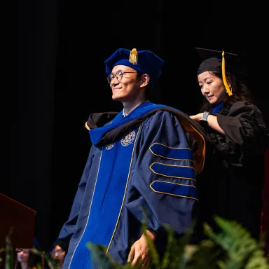 A young man with glasses wearing a blue graduation robe with a doctoral hood being placed around his shoulders with a woman standing behind him