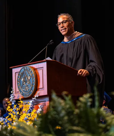 A man with glasses wearing a graduation robe standing a podium with the Drexel University seal