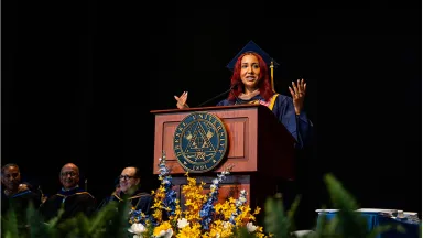 A young woman with red hair wearing a graduation robe and cap standing at a podium gesturing with both hands