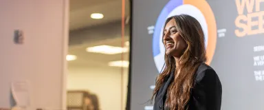 Woman with long brown hair in black blazer standing in front of projector screen.