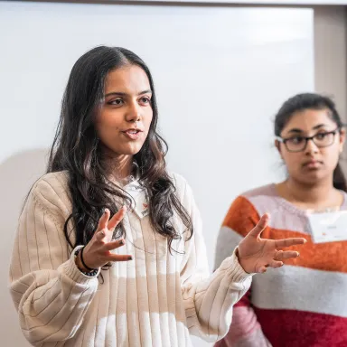 Woman speaking in front of a projector screen with another woman behind her.