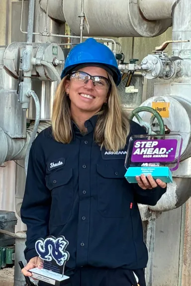 Woman in blue hard hat and work uniform holding two awards in an industrial setting