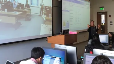 Students in classroom with a professor at the front and another class of students visible on a projector screen