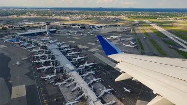 An overhead view of numerous airplanes lined up at an airport, with the wing of an airborne plane visible in the lower right