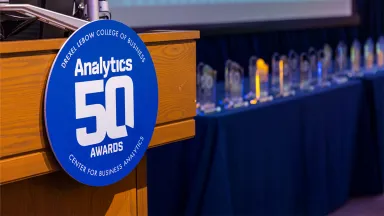 A circular blue badge with the text Analytics 50 Awards on a wooden podium, with clear glass awards visible in the background