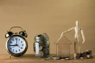 A clock sits next to a jar filled with coins. A wooden figure stands nearby with one hand on a wooden house, coins stacked around it.