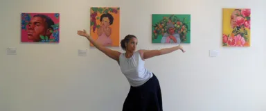 A young woman in a white top and black skirt with arms extended, standing in front of colorful artwork display on a white gallery wall