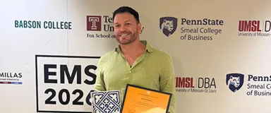 A young man wearing a green shirt holding an orange certificate and standing in front of a backdrop with multiple university and corporate logos