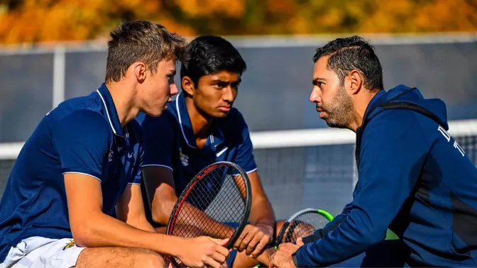 Two young men wearing navy polo shirts facing and talking to an older man wearing a navy jacket