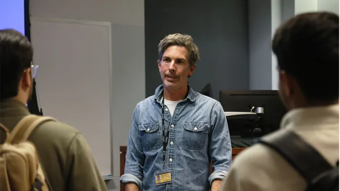Man wearing denim shirt and a lanyard speaking to two students with their backs to the camera