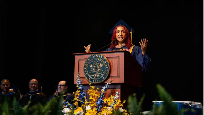 A young woman with red hair wearing a graduation robe and cap standing at a podium gesturing with both hands