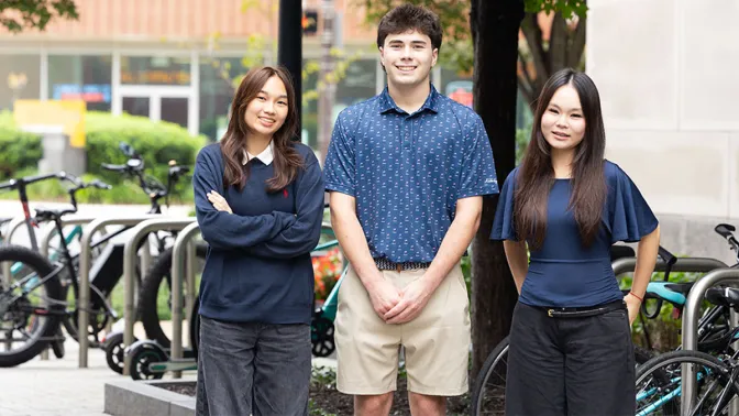 Three individuals - a young woman, a young man, and another young woman - in casual attire with a tree and several bicycles visible in the background