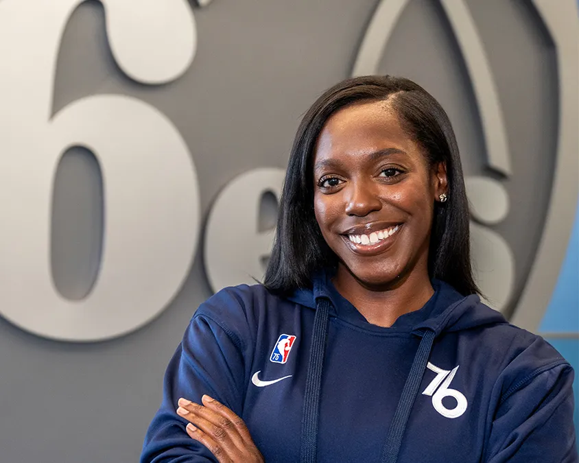 A young Black woman in a blue sweatshirt with the Philadelphia 76ers logo, standing in front of a large silver rendition of the 76ers logo and with her arms crossed in front of her chest