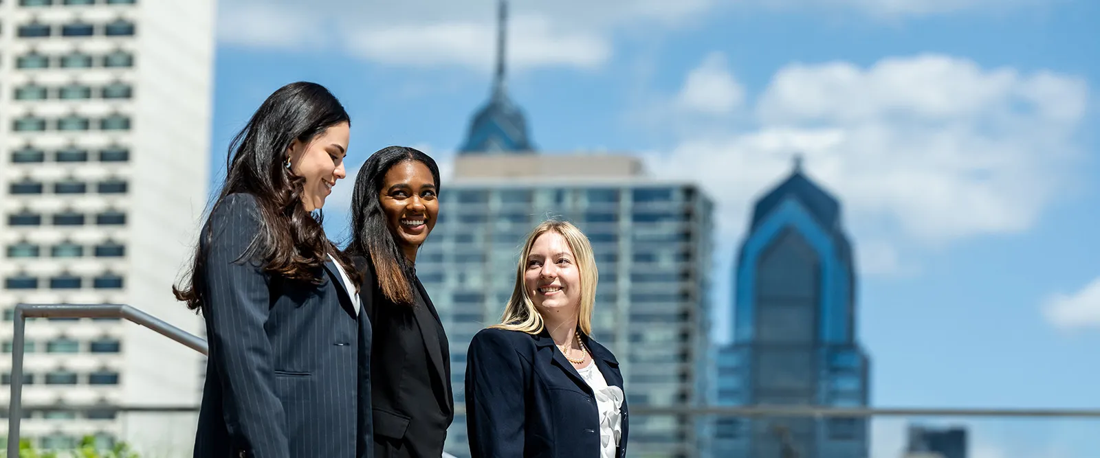 three women in business attire walking on lebow's campus with iconic philadelphia buildings visible in the background